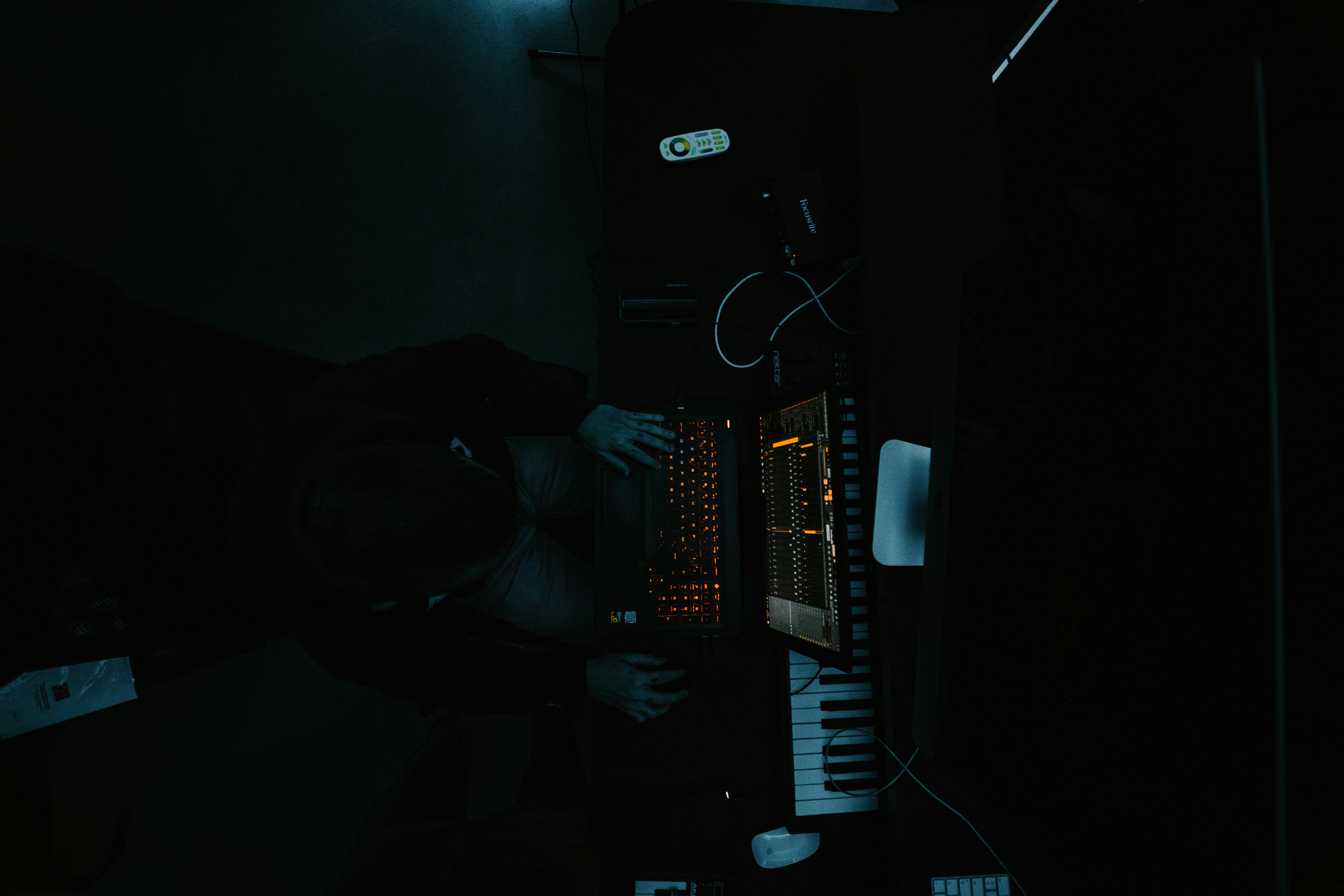 A producer sits in a dimly lit music studio, working on a laptop with audio equipment.