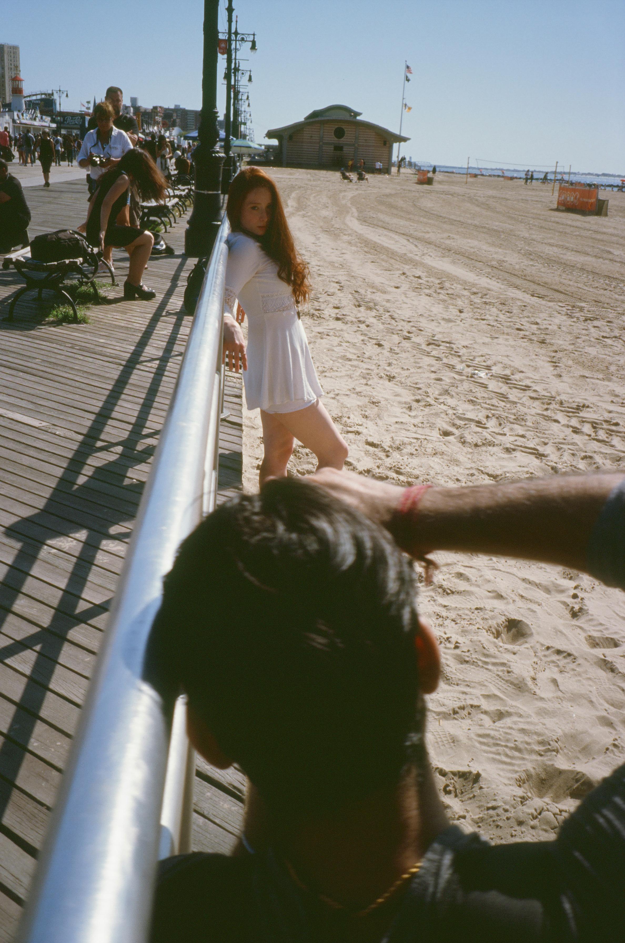 Home Fashion photoshoot on a sunny beach boardwalk featuring a model in a white dress, capturing stylish summer vibes.