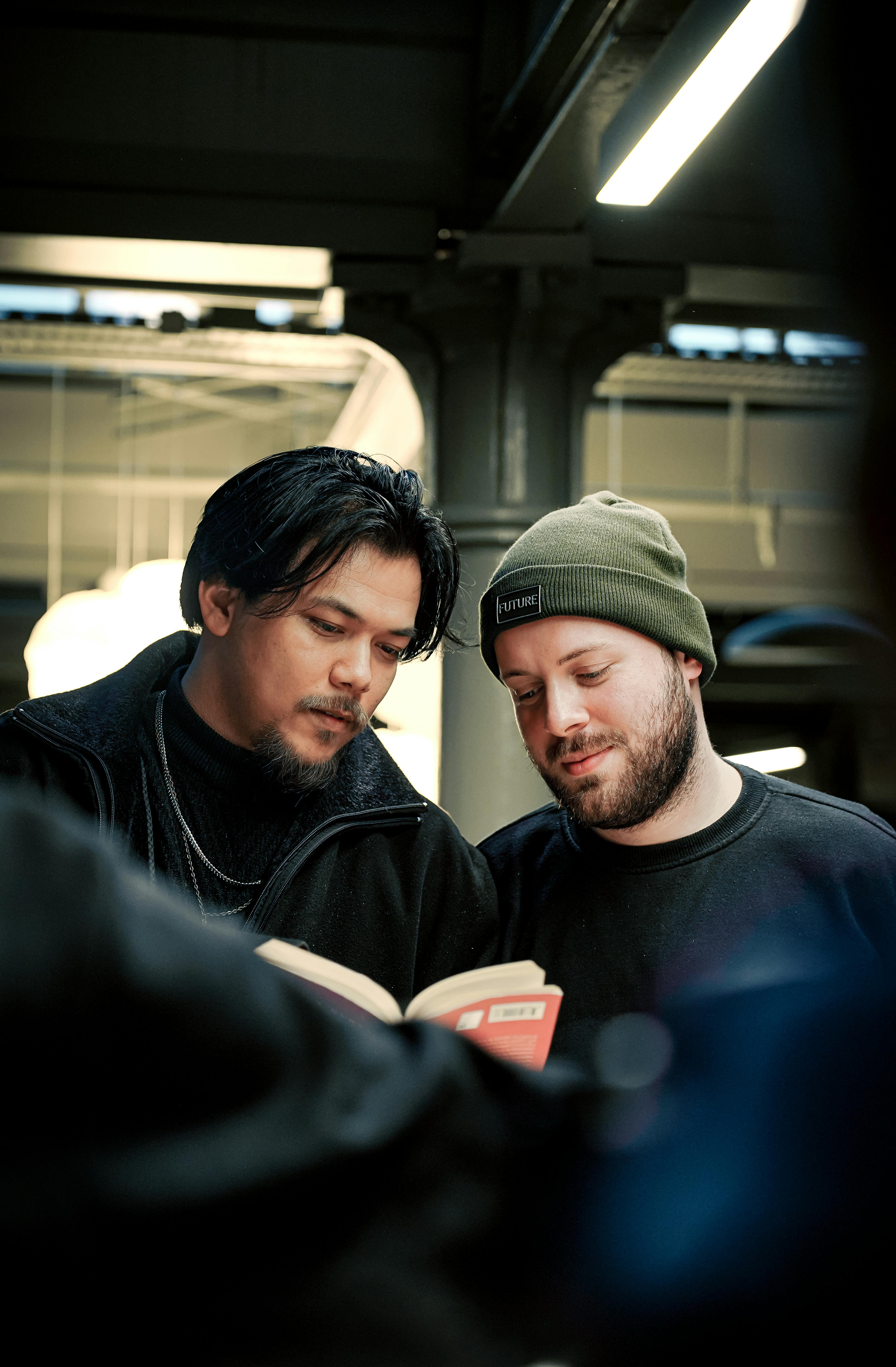 Two men engaged in reading a book together in a cozy indoor environment.