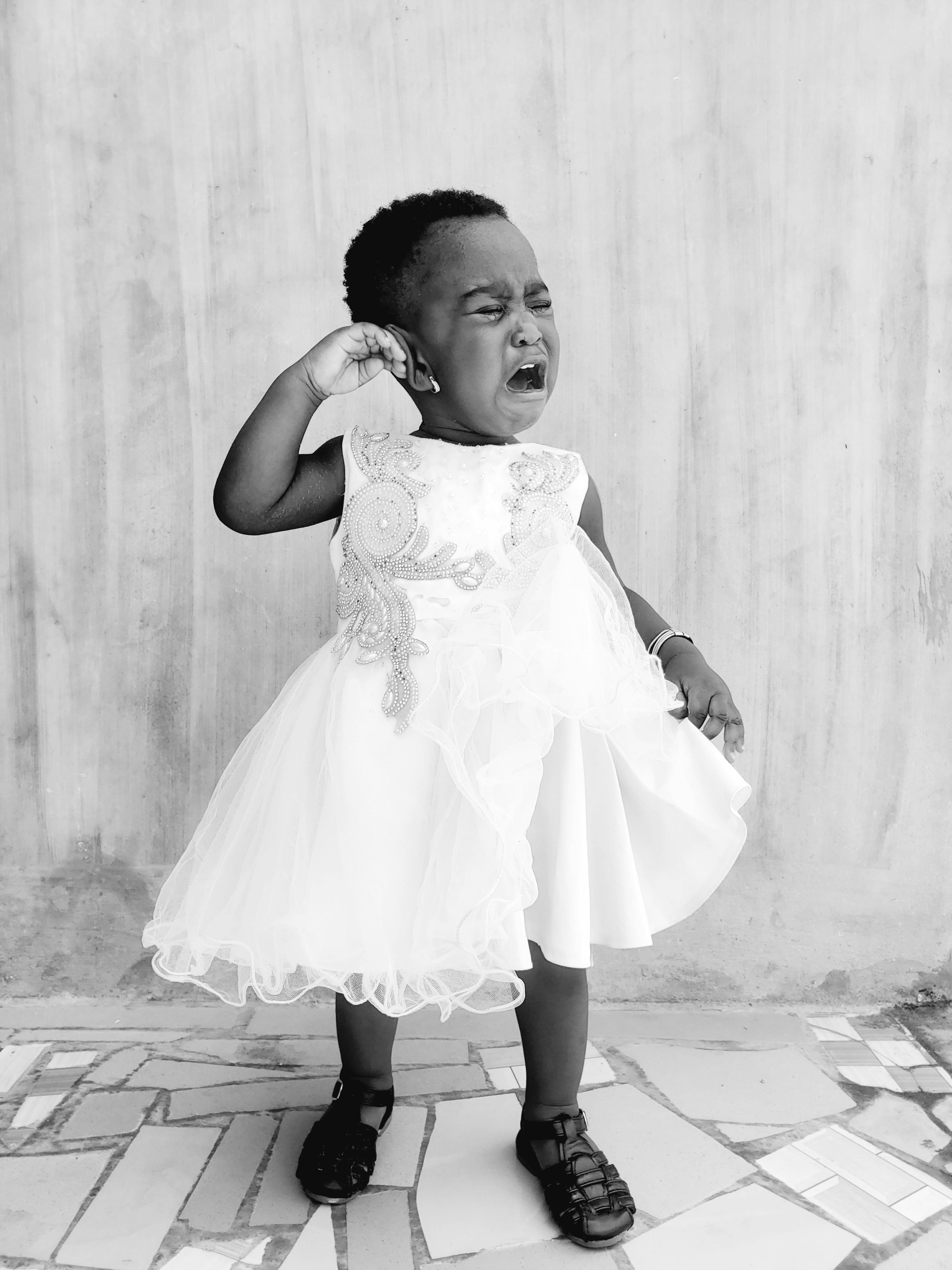 Black and white portrait of a crying child in a white dress standing indoors, expressing emotion.
