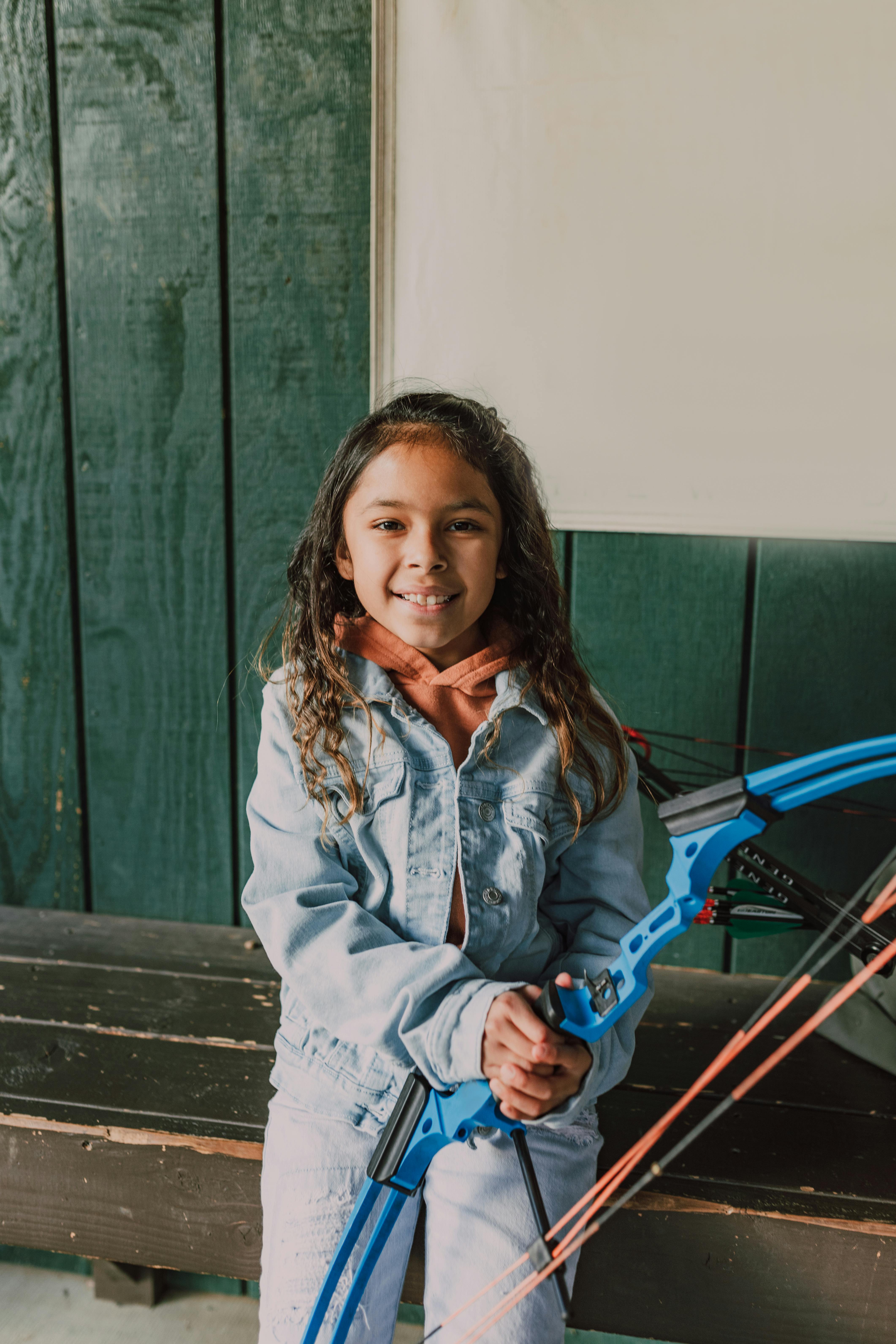 Services A young girl with a bow stands indoors during archery practice, showcasing focus and skill.