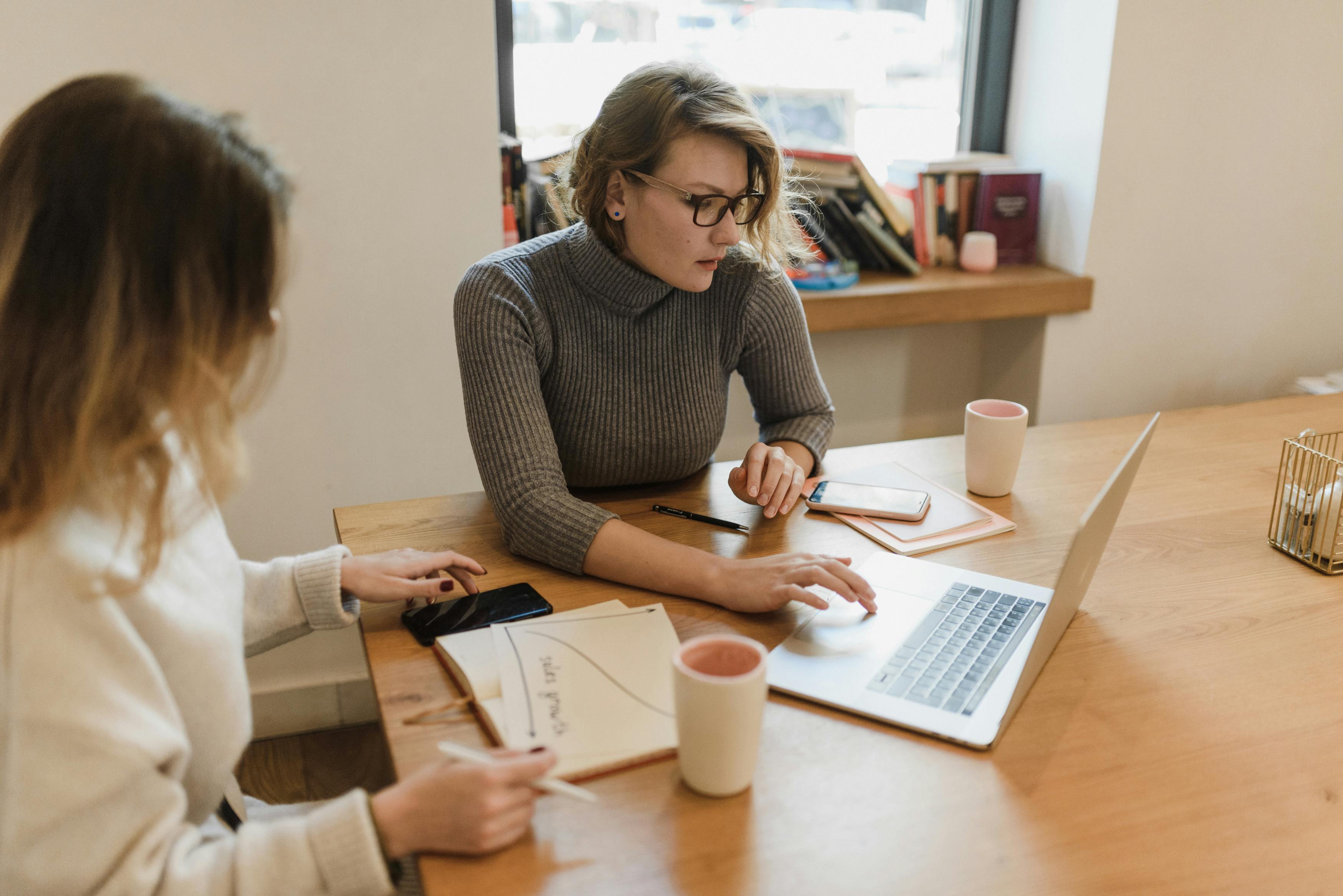 Two women working together with a laptop and notebooks in an office setting.