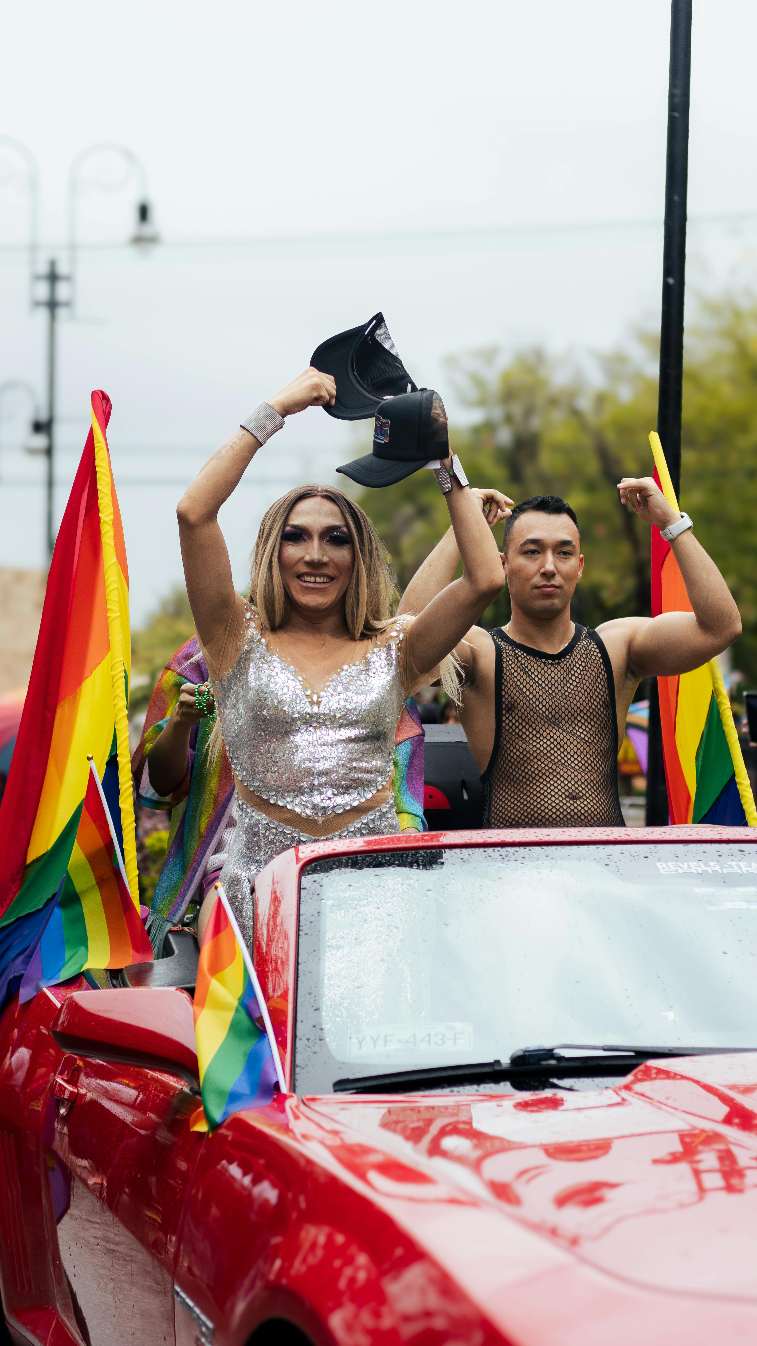 Vibrant pride parade scene with a red convertible and lively participants waving rainbow flags.