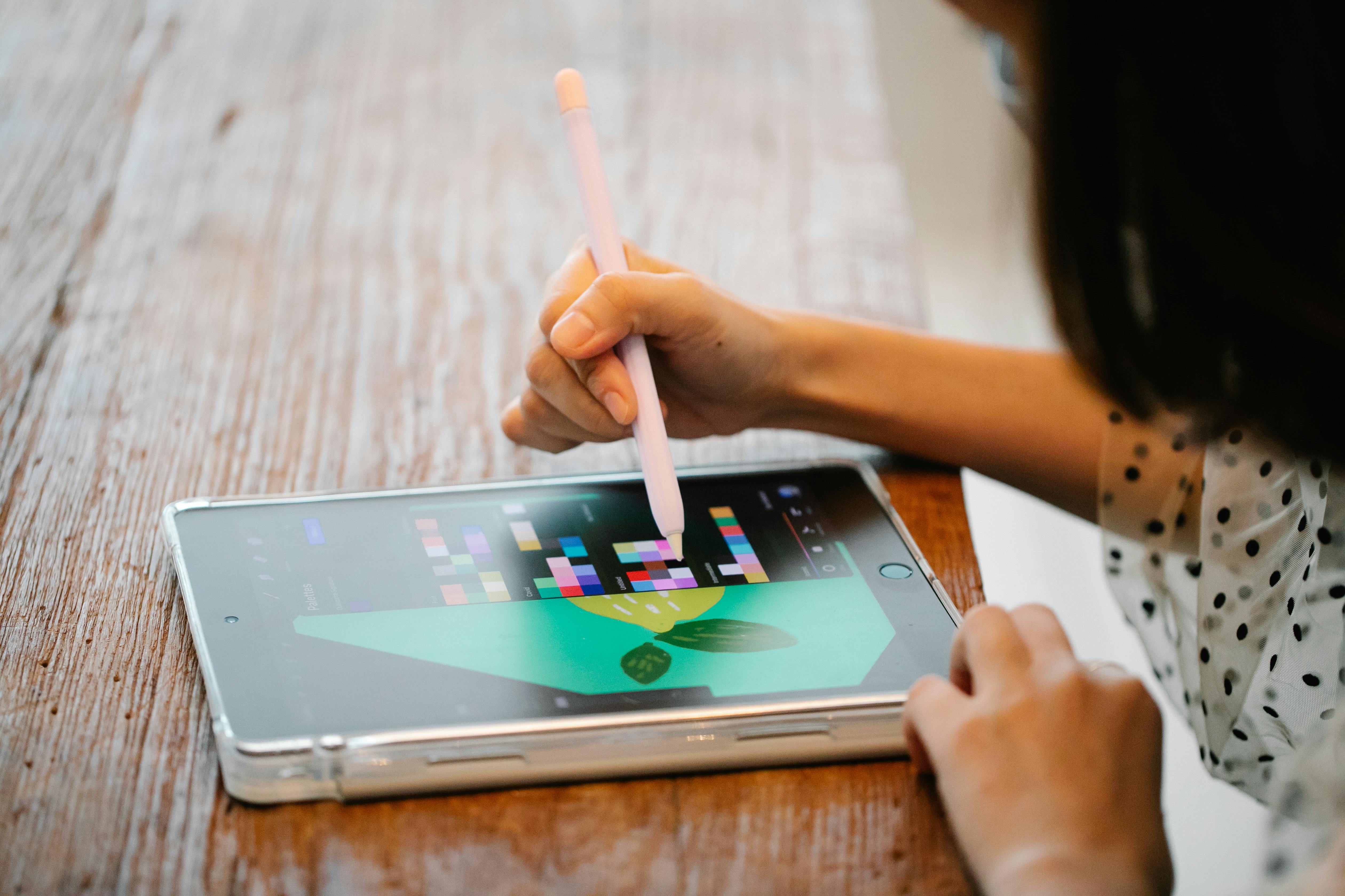 A person using a stylus pen on a tablet for digital illustration at a wooden desk.